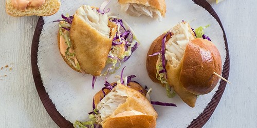 Mini fish sliders with flaky hoki fillets, shredded cabbage, and sauce in soft golden buns, served on a white plate.