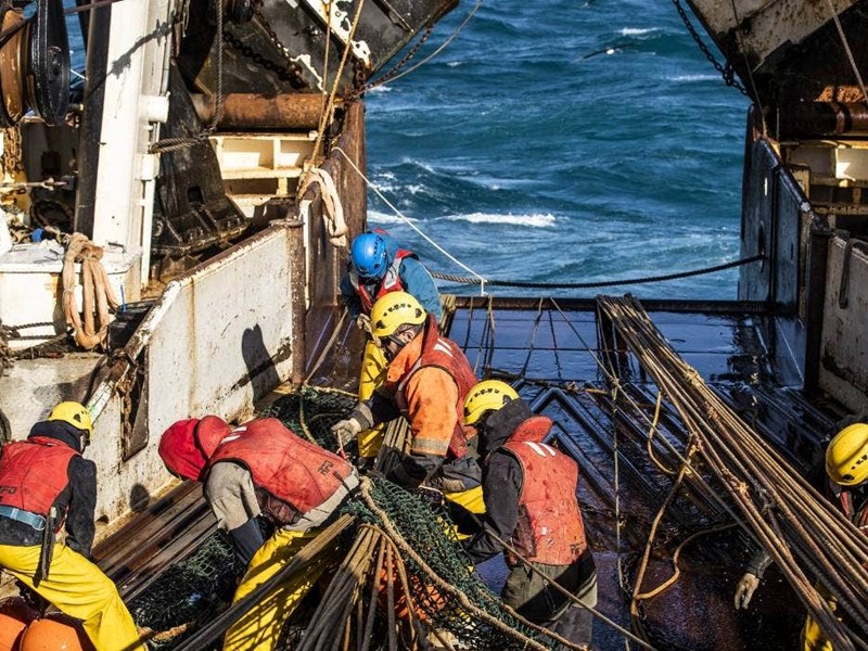 Crew members wearing safety gear working with fishing nets on the deck of a commercial fishing vessel at sea.