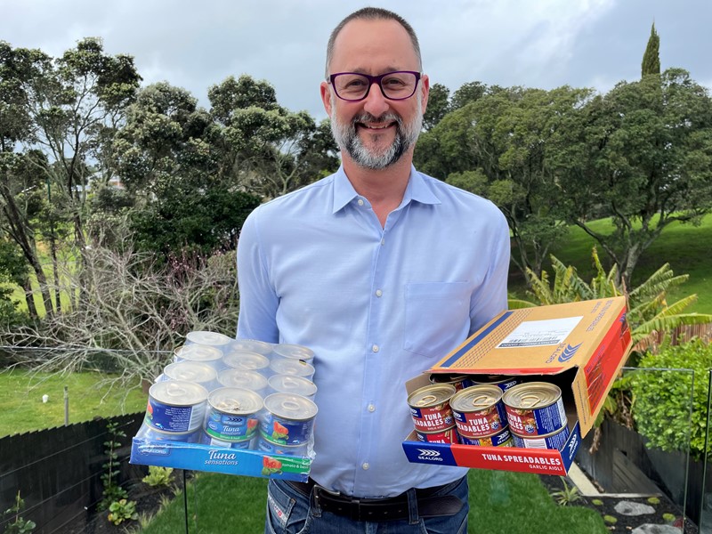 Man wearing glasses and a blue button-up shirt holding two trays of canned tuna, standing in front of a lush green garden with trees in the background.