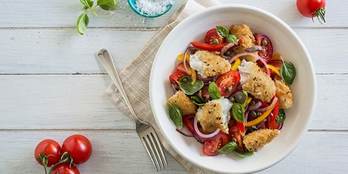 Tuscan-style fish salad with crispy fish pieces, cherry tomatoes, olives, basil, red onion, and capsicum—served in a white bowl on a rustic wooden table.