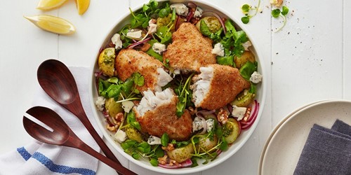 A colourful winter fish salad featuring golden pan-seared fish portions atop a bed of leafy greens, roasted Brussels sprouts, walnuts, red onion, and blue cheese.