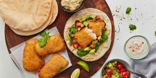 Flatbread topped with crumbed hoki fillet, avocado salsa, spinach, and hummus, surrounded by extra flatbreads, sauces, and garnishes.