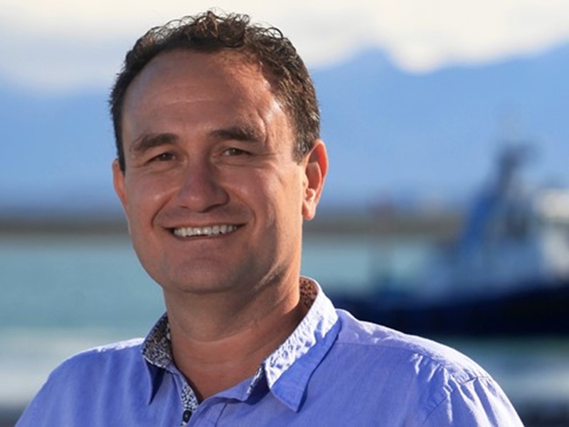 Sealord Chief Executive Officer smiling at the camera with the ocean, distant Sealord vessel, and mountains in the background.