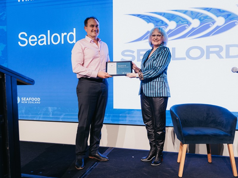 Two people standing on stage holding the Future Adaptation Award, with the blue and white Sealord logo displayed in the background, celebrating Sealord’s recognition at the 2024 Seafood Star Awards for its significant sustainability efforts.