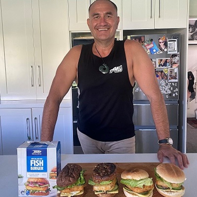 Man smiling in a kitchen standing behind a row of homemade fish burgers made with Sealord New Zealand Hoki Fish Burger patties
