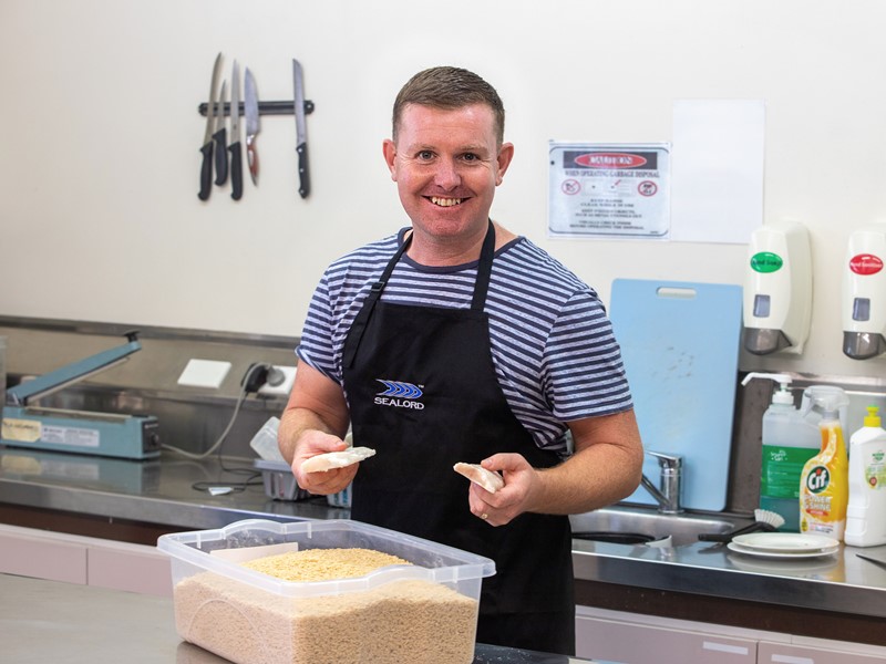 Smiling man in a Sealord apron breading fish fillets in a commercial kitchen, holding two pieces above a container of breadcrumbs.