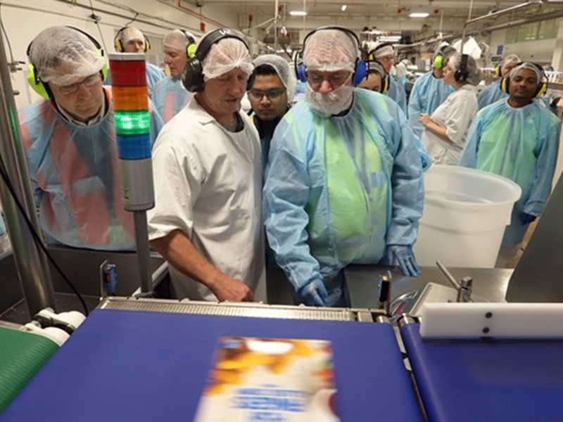 Factory line workers wearing safety gear, including gloves, hairnets, and earmuffs, inspecting a box of frozen fish as it moves along a conveyor belt.