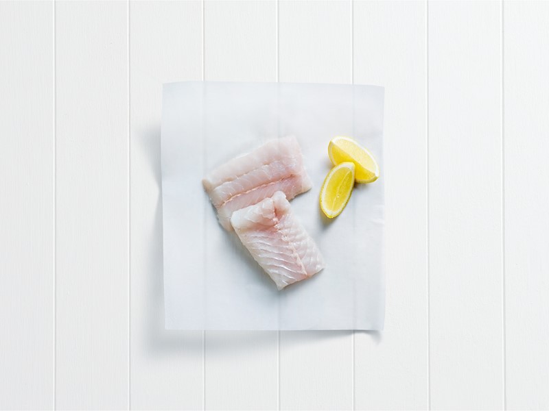 Two raw white fish fillets and lemon wedges placed on white baking paper over a white wooden surface
