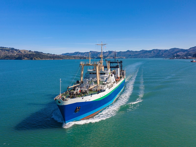 Large commercial fishing vessel sailing through calm turquoise waters with hills and coastline in the background under a clear blue sky.