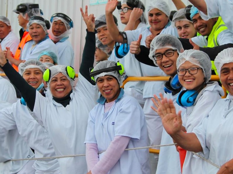 Sealord staff smiling and waving at the camera, wearing factory uniforms, hair nets, and earmuffs outside the Sealord processing facility.
