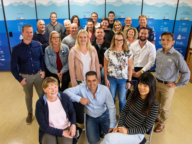 Group of office workers posing together and smiling for a team photo indoors, with an illustrated ocean and ship-themed backdrop behind them.