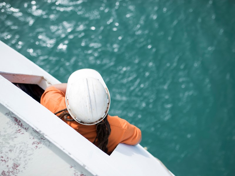 birds eye shot of a man leaning out of a sealords vessel window looking down at the sea he is wearing orange overalls and a white safety hard hat.
