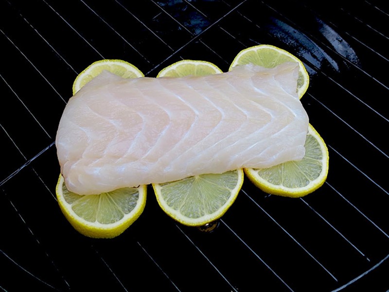 Raw white fish fillet placed on a bed of lemon slices on a grill.