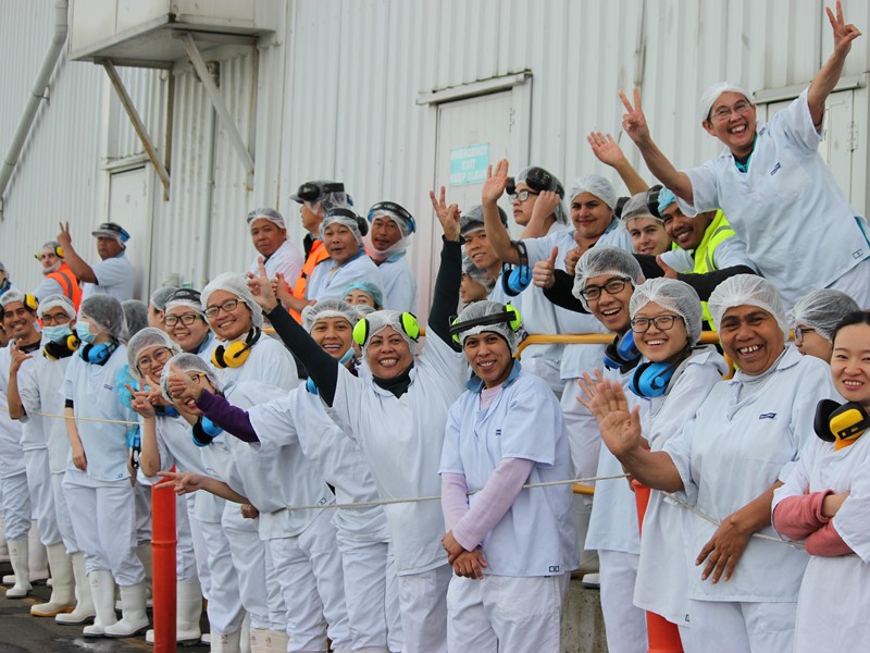 A cheerful group of seafood processing workers dressed in white uniforms and safety gear, smiling and waving outside a factory building.