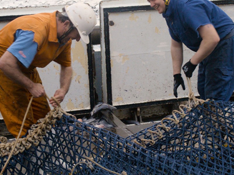 two men tying up a fishing net together, one man is wearing orange overalls and a white saftey and the other is wearing blue overalls