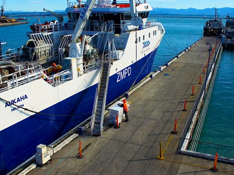 Sealord fishing vessel named AUKAHA docked at a port in Nelson, New Zealand. A worker in high-visibility gear stands on the wharf beside stacked crates, with clear blue water and mountains in the background.