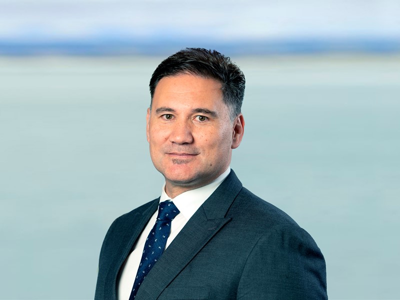 Man wearing a dark suit, white shirt, and navy tie with small patterns, standing in front of a soft ocean and horizon backdrop.