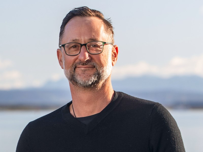 Smiling man with short dark hair, glasses, and a salt-and-pepper beard, wearing a black sweater, standing in front of a scenic ocean and mountain backdrop