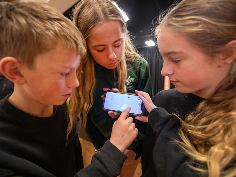 three children huddled around holding a phone in landscape view showing the Sealord is launching its updated educational fishing game app