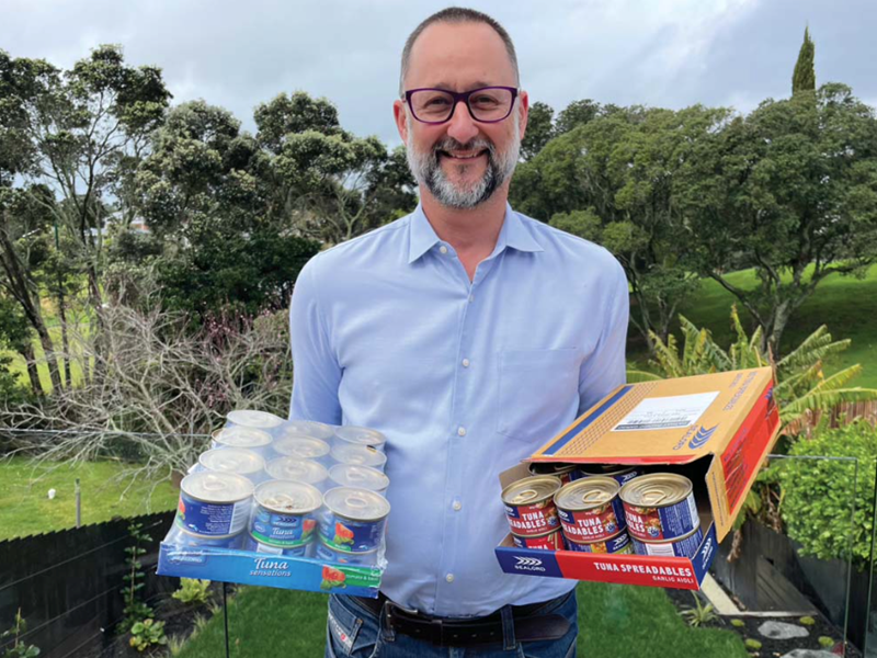 Man wearing glasses and a blue button-up shirt holding two trays of canned tuna, standing in front of a lush green garden with trees in the background.
