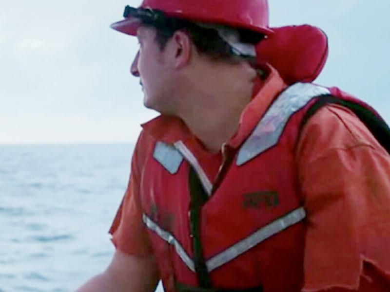 Person wearing a red hard hat, orange safety vest, and work shirt, looking out over the ocean. Appears to be involved in marine or fishing operations.