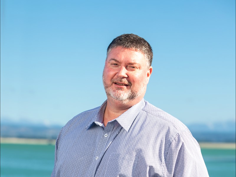 Smiling man with short dark hair and a trimmed beard, wearing a light blue checkered shirt, standing in front of a bright blue sky and ocean backdrop
