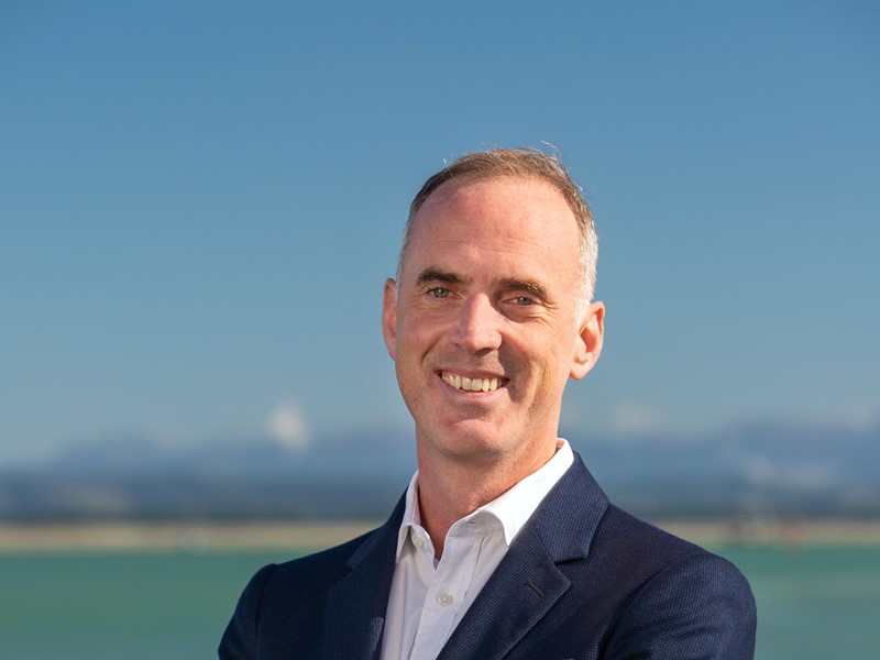 "Smiling man with short light brown hair wearing a white shirt and navy blazer, standing in front of a bright ocean and mountain backdrop