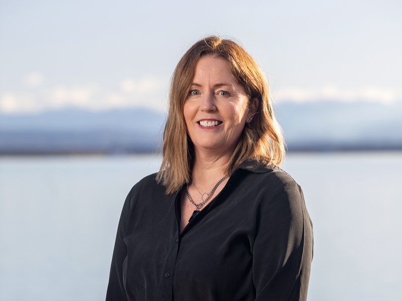 Woman with shoulder-length light brown hair wearing a black shirt, smiling in front of a scenic water and mountain background