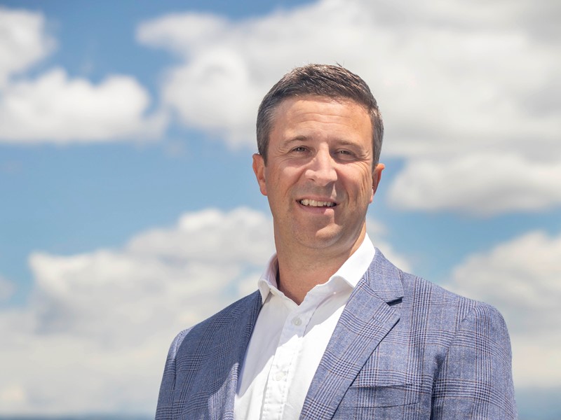 Smiling man wearing a light blue checked blazer and white shirt, standing outdoors against a bright blue sky with scattered clouds