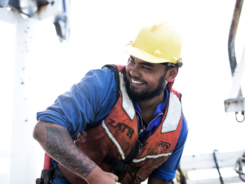 sealord worker smiling looking towards his left, he is wearing a blue shirt, orange life Jacket and yellow hard hat