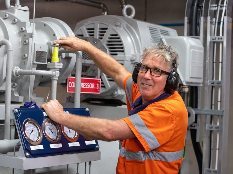 Worker in orange safety gear adjusting machinery gauges in an industrial setting, wearing hearing protection and glasses