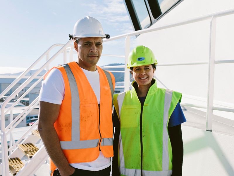 Two workers wearing safety vests and helmets standing on the deck of a ship, smiling in bright daylight.