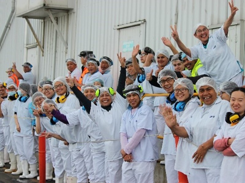 Large group of smiling factory workers wearing white uniforms, hairnets, and ear protection, gathered outside a building and waving at the camera