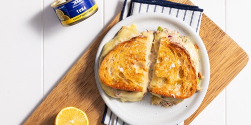 Cracked pepper tuna toastie from Sealord’s recipe range, cut in half and served on a white plate atop a wooden chopping board, with half a lemon in the bottom left and a can of Sealord tuna in the top left.
