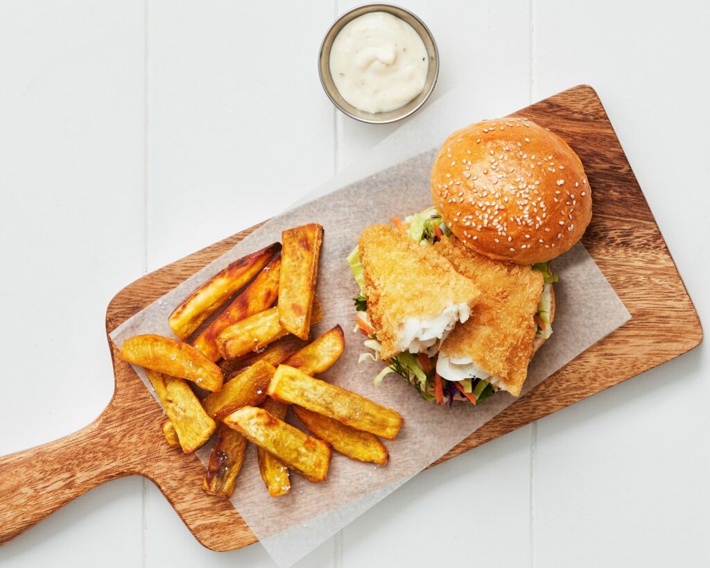 Crispy Hoki Panko fish burger with slaw on a sesame seed bun, served with thick-cut golden fries and dipping sauce on a wooden board.
