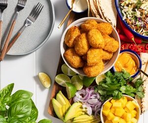 Platter on wooden board featuring Sealord fish bites, avocado, lime, mango, chilli, coleslaw, red onion, and soft tacos, with an empty plate and three forks on the left side.