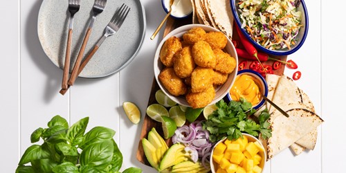 Platter on wooden board featuring Sealord fish bites, avocado, lime, mango, chilli, coleslaw, red onion, and soft tacos, with an empty plate and three forks on the left side.