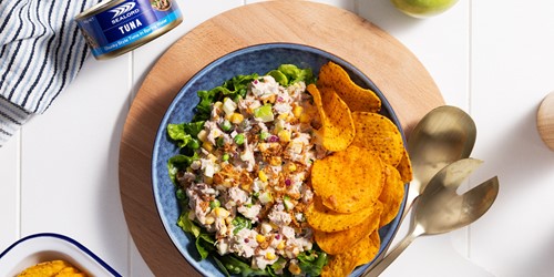 Bowl of creamy tuna salad with chopped vegetables served on lettuce, paired with crunchy seasoned chips. Set on a wooden board with gold salad servers, a can of Sealord tuna, and a striped cloth in the background on a white tabletop.