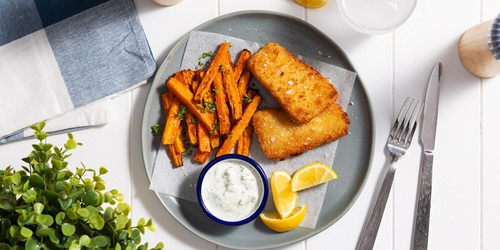 Plate with two golden crumbed fish fillets, seasoned carrot fries, a small bowl of tartar sauce, and lemon wedges. Set on a grey plate with parchment paper, surrounded by cutlery, a glass, a napkin, and greenery on a white tabletop.