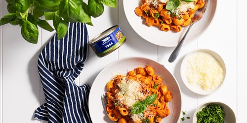 Two bowls of pasta in a rich tomato sauce topped with grated cheese and fresh basil, accompanied by a can of Sealord tuna, a bowl of grated cheese, chopped herbs, and a navy-striped napkin on a white tabletop. Fresh basil leaves hang above the scene