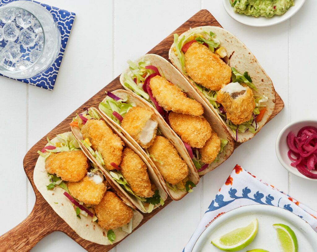 Tacos filled with crispy Hoki Bites, shredded lettuce, slaw, and pickled onions, served on a wooden board with sides of guacamole, lime, and iced water.