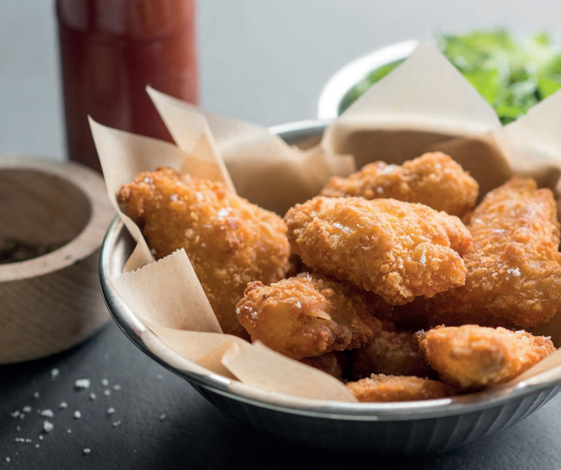 Bowl lined with parchment paper filled with golden, crispy battered fish bites. Surrounded by a jar of sauce, a wooden bowl with seasoning, and fresh herbs in the background.