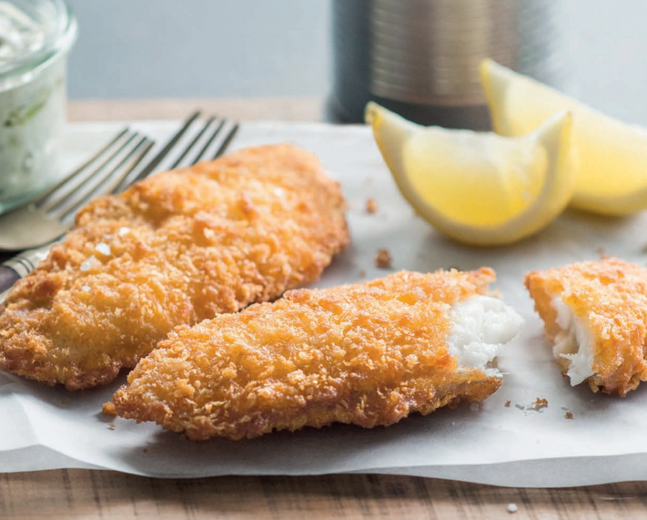 Two golden, crispy battered fish fillets served on parchment paper with lemon wedges, a broken piece revealing flaky white fish inside, and forks placed in the background.