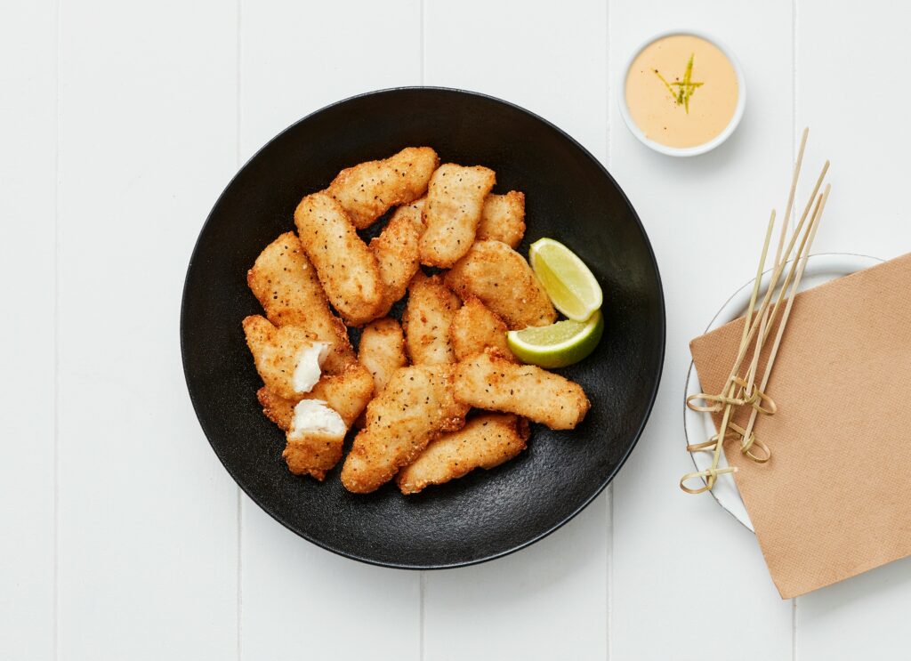 Crispy Hoki salt and pepper fillet bites served in a black bowl with lime wedges, dipping sauce, and skewers on the side.