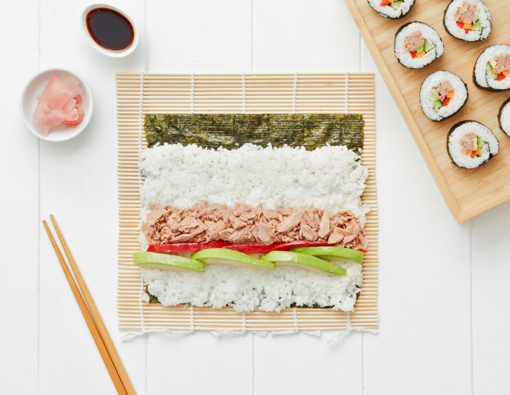 Sushi roll being made with tuna flakes, avocado, capsicum, and rice on a bamboo mat, with finished rolls and condiments nearby.