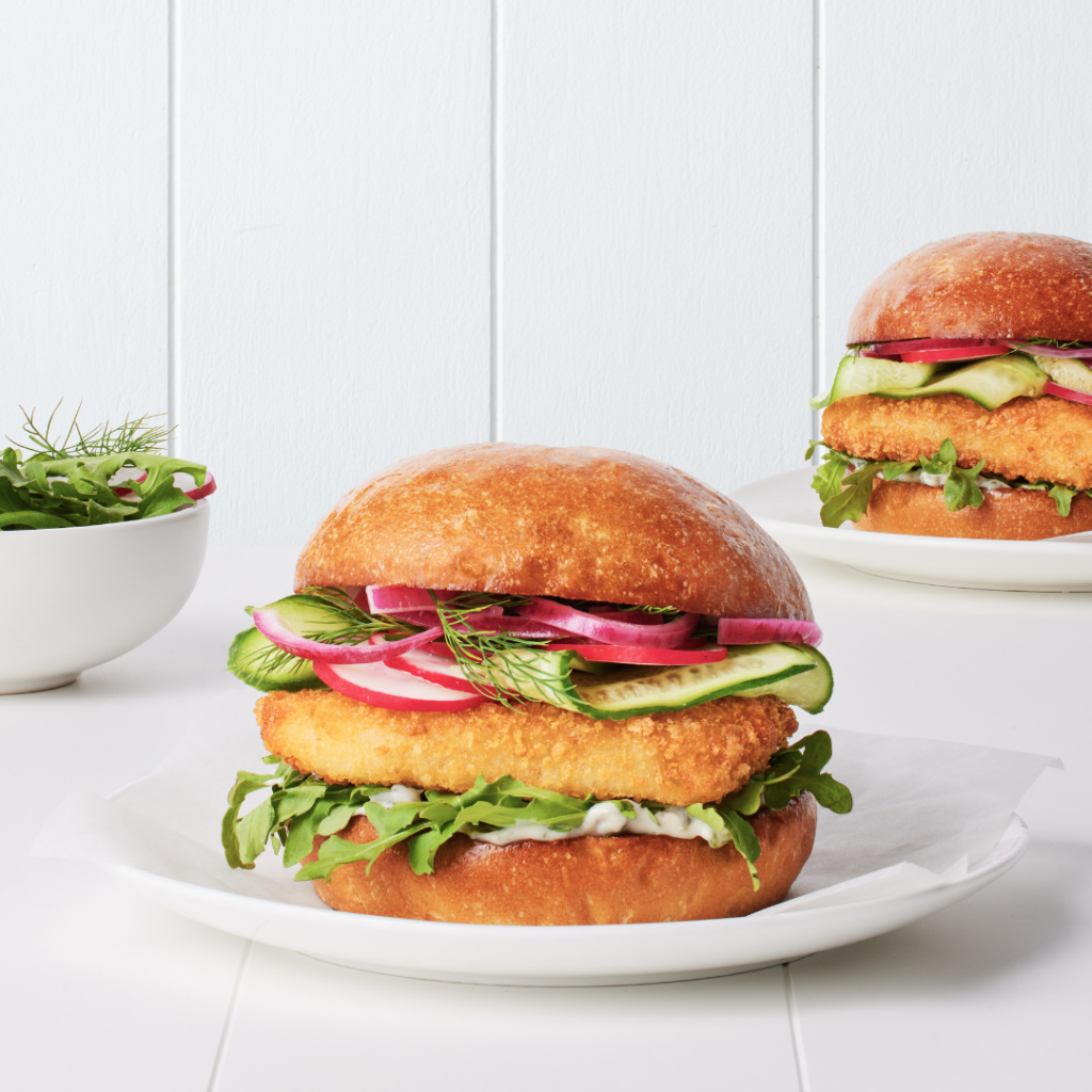Crispy hoki burger on a toasted bun with fresh rocket, tartare sauce, pickled onions, cucumber ribbons, and dill – served on a white plate.
