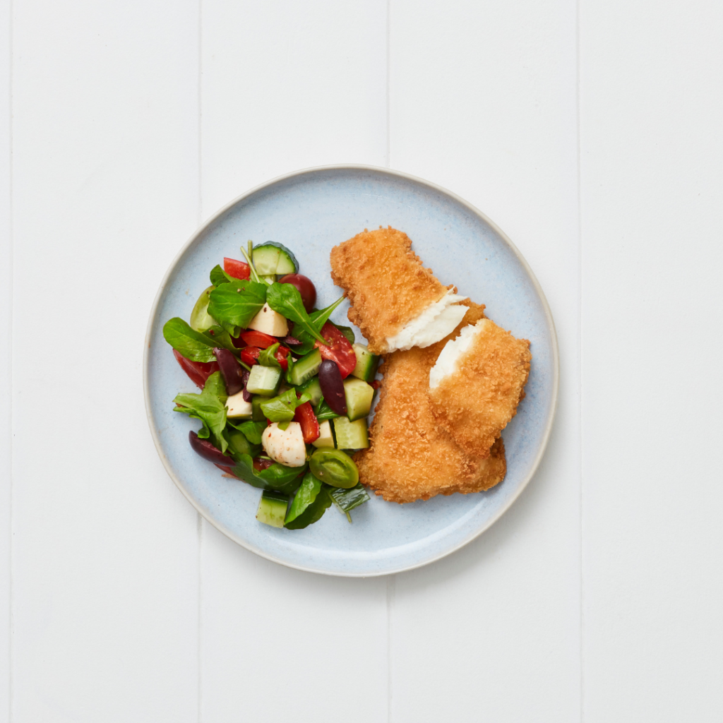 Gluten-free crumbed Hoki portions served on a plate with a fresh salad of cucumber, cherry tomatoes, olives, capsicum, and mozzarella.